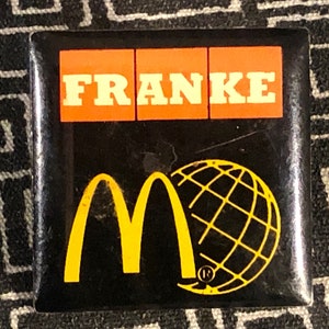 May include: A black and yellow pin with the McDonald's golden arches logo and a globe with the word "FRANKE" in orange letters at the top.