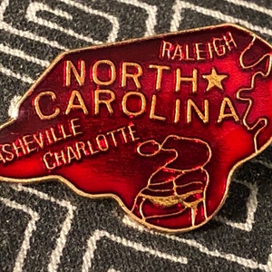 Puede incluir: Un pin de esmalte rojo con forma del estado de Carolina del Norte con las palabras "North Carolina" y "Raleigh" en letras doradas. El pin tambi&eacute;n incluye las ciudades de "Asheville" y "Charlotte" en letras doradas.