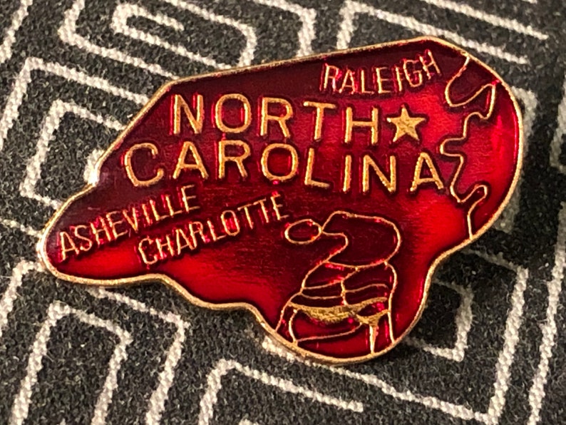 Puede incluir: Un pin de esmalte rojo con forma del estado de Carolina del Norte con las palabras "North Carolina" y "Raleigh" en letras doradas. El pin tambi&eacute;n incluye las ciudades de "Asheville" y "Charlotte" en letras doradas.