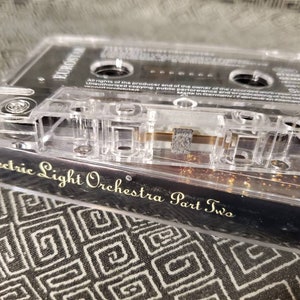 May include: A clear cassette tape case with a black cassette tape inside. The cassette tape is labeled "Electric Light Orchestra Part Two" in gold lettering. The case has a barcode and the text "Side 2" printed on it.