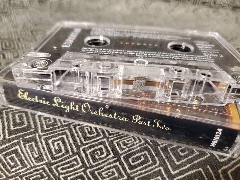 May include: A clear cassette tape case with a black cassette tape inside. The cassette tape is labeled "Electric Light Orchestra Part Two" in gold lettering. The case has a barcode and the text "Side 2" printed on it.