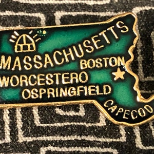 May include: A gold-colored pin shaped like the state of Massachusetts. The pin is green with a gold outline and features the words "Massachusetts", "Worcester", "Boston", "Springfield", and "Cape Cod".