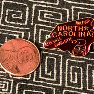 Puede incluir: Un pin de esmalte rojo y dorado con forma del estado de Carolina del Norte. El pin dice "Carolina del Norte" con las ciudades "Raleigh", "Asheville" y "Charlotte" enumeradas. Hay una estrella dorada en el pin.