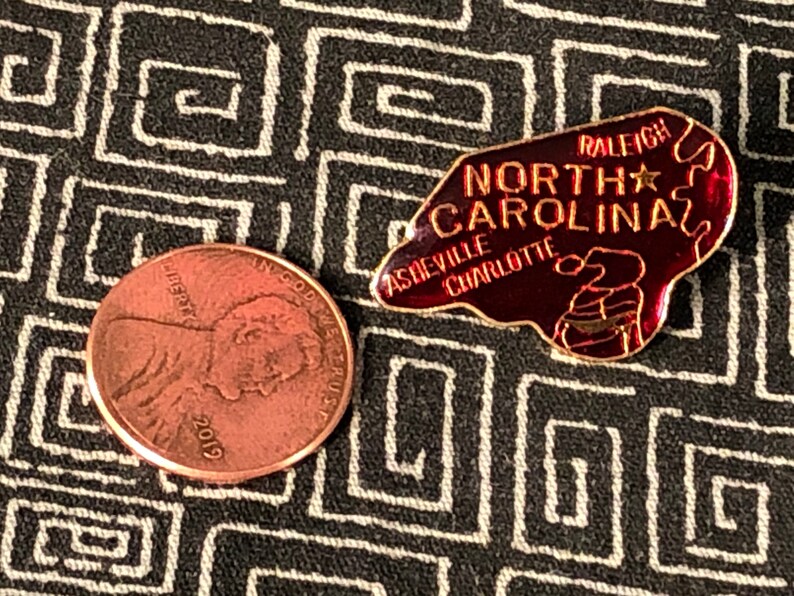 Puede incluir: Un pin de esmalte rojo y dorado con forma del estado de Carolina del Norte. El pin dice "Carolina del Norte" con las ciudades "Raleigh", "Asheville" y "Charlotte" enumeradas. Hay una estrella dorada en el pin.