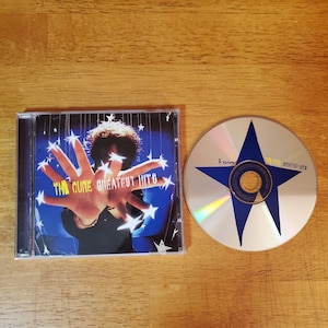 May include: A The Cure Greatest Hits CD in a clear plastic jewel case with a cover featuring a person with outstretched hands and white star graphics, alongside the metallic disc which displays a large blue star and the text The Cure Greatest Hits.