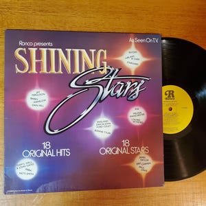 May include: A vintage vinyl record album titled "Shining Stars" by Ronco, featuring 18 original hits and stars. The album cover is purple and features the album title in a stylized font. A black vinyl record is next to the album.