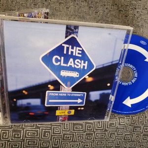 May include: A CD case with a blue and white design featuring a road sign that says "The Clash" and "From Here To Eternity". The CD case is open and the CD is visible. The CD is blue with a white arrow.