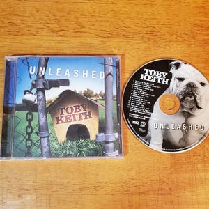 May include: A Toby Keith Unleashed CD in a clear plastic case with a cover showing a doghouse behind a chain-link fence and a padlock. The disc features a black and white photograph of a bulldog and a track list printed in white text.