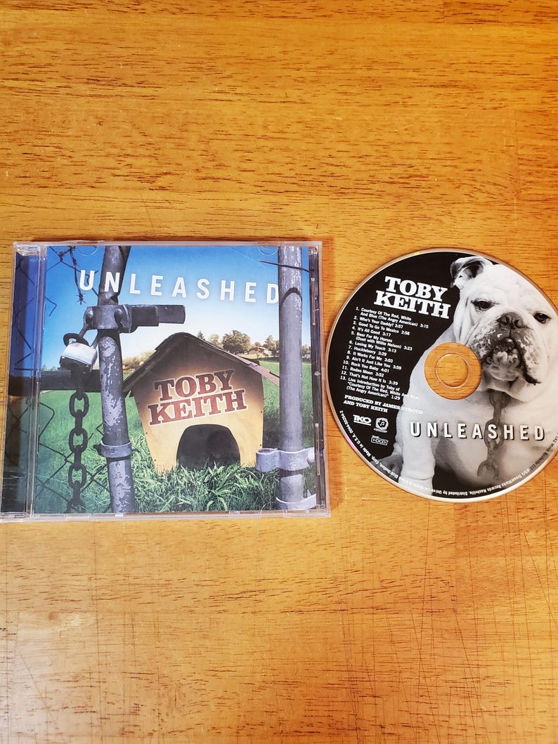 May include: A Toby Keith Unleashed CD in a clear plastic case with a cover showing a doghouse behind a chain-link fence and a padlock. The disc features a black and white photograph of a bulldog and a track list printed in white text.
