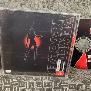 May include: A CD case and disc for the album "Contraband" by Velvet Revolver. The case features a black background with a silhouette of a woman holding a gun, and the band's name in white. The disc has a red and black design.