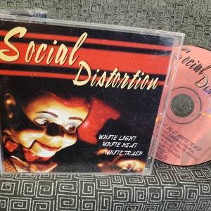 May include: A CD case with a red and black background featuring a puppet with a white face and red lips. The text "Social Distortion" is written in yellow on the front of the case. The CD is visible in the background with the text "Social Distortion" written in black on a pink background.
