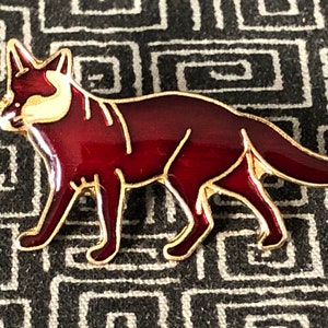 May include: A red enamel fox pin with gold accents. The fox is depicted in a walking pose with its tail curled.