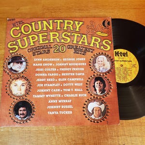 May include: A K-Tel Country Superstars vinyl record album cover featuring a brown background with yellow text, 20 greatest hits, and portraits of country artists. The record is black with a yellow K-Tel label, showing track listings in black text.