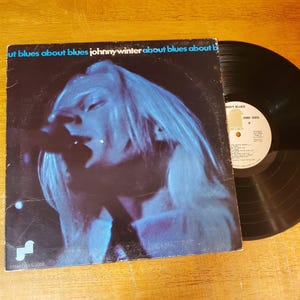 May include: A vintage vinyl record album by Johnny Winter, titled "About Blues About Blues." The album cover features a blue-toned image of Johnny Winter singing. The record is black and sits on a wooden surface.