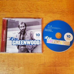 May include: A Lee Greenwood 10 Great Songs music CD in a clear plastic jewel case with a black and white cover photo of the artist. The disc is solid blue with white text and a matching 10 Great Songs logo. The artist name is printed in blue.