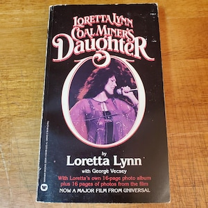 May include: A vintage paperback book titled "Loretta Lynn Coal Miner's Daughter" with a photo of Loretta Lynn on the cover. The cover is black with white and red text. The book includes a photo album and is now a major film.