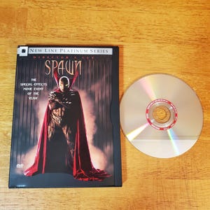 May include: A Spawn Director's Cut DVD from the New Line Platinum Series. The black case features the character Spawn in a red cape and the text The Special-Effects Movie Event of the Year. A silver DVD disc is positioned to the right of the case.