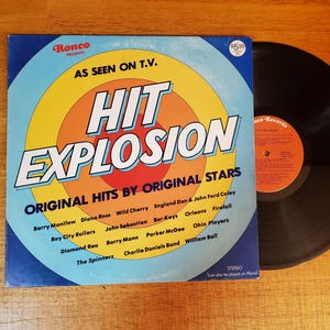 May include: A vintage Hit Explosion vinyl record album cover featuring a blue, yellow, and orange circular graphic design. The text reads Ronco Presents, As Seen On T.V., Hit Explosion, and Original Hits By Original Stars, with a list of artists.