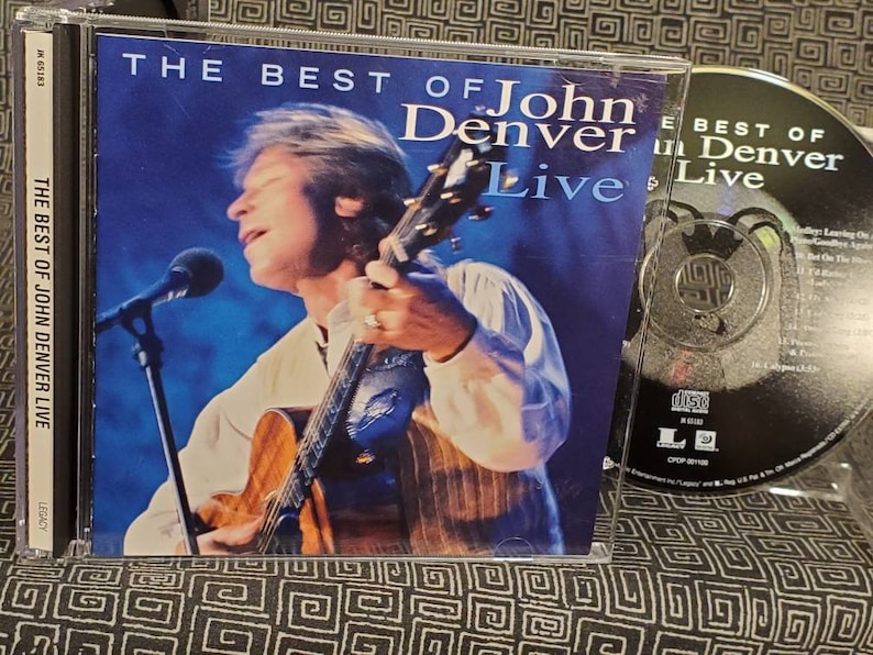 May include: A CD case with a blue background featuring a photo of John Denver playing a guitar. The text on the case reads "The Best of John Denver Live".