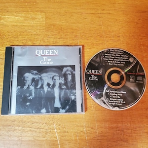 May include: A CD and jewel case for Queen's album "The Game" are displayed on a wooden surface. The CD is visible next to the case, with the album title and tracklist. The album cover features a black and white photo of the band.