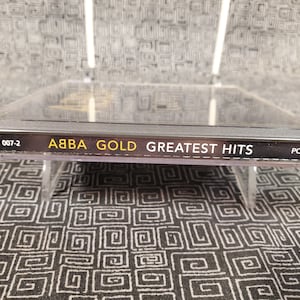 May include: ABBA Gold: Greatest Hits CD. The CD case is clear, with the album title and artist name in gold and white lettering. The CD is displayed on a clear stand against a patterned background.