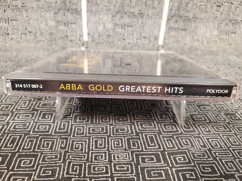 May include: ABBA Gold: Greatest Hits CD. The CD case is clear, with the album title and artist name in gold and white lettering. The CD is displayed on a clear stand against a patterned background.