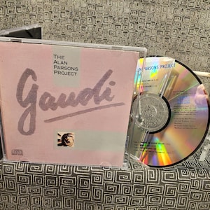 May include: A pink CD case with the title "The Alan Parsons Project Gaudi" printed on the front. The CD case is next to a silver CD with a rainbow holographic design.