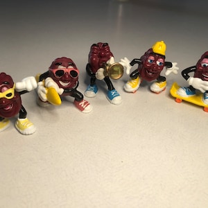 May include: Five small plastic figurines of red grapes with different accessories. One grape is wearing sunglasses and holding a banana, another is wearing a yellow hard hat and holding a skateboard, another is wearing sunglasses and holding a trumpet, another is wearing sunglasses and holding a boombox, and the last one is wearing sunglasses and holding a skateboard.