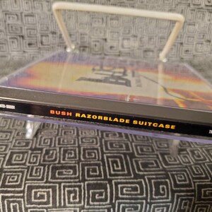 以下が含まれることがあります： 黄色とオレンジの背景を持つ黒いCDケース。ケースの前面には「Bush Razorblade Suitcase」という文字が印刷されています。ケースの右側には「Trauma Records」という文字が印刷されています。