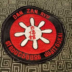 May include: A black and red round patch with white embroidery. The patch features a stylized wheel with eight spokes and Japanese characters in the center. The text "DAN ZAN RYU STROUDSBURG JU-JITSU-KAI" is embroidered around the wheel.