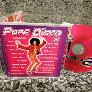May include: A pink CD case with a purple and white background. The case has the title "Pure Disco 2" and lists artists such as Gloria Gaynor, ABBA, Van McCoy, and Diana Ross. The CD is pink and has the title "Pure Disco 2" on it.