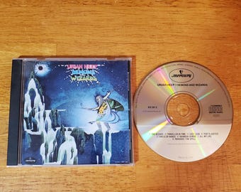 May include: A compact disc and its plastic jewel case featuring the album Demons and Wizards by Uriah Heep. The cover art displays a blue landscape with a figure, while the silver disc lists track titles including The Wizard and Easy Livin.