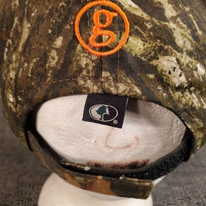 Peut inclure: Une casquette de baseball camouflage avec un logo "G" orange sur le devant et un patch noir avec un logo d'arbre au dos.