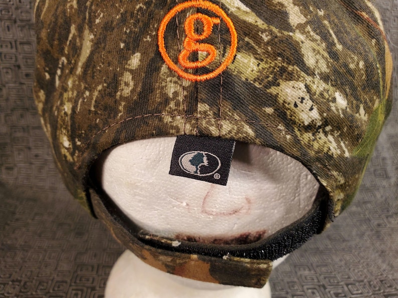 Peut inclure: Une casquette de baseball camouflage avec un logo "G" orange sur le devant et un patch noir avec un logo d'arbre au dos.