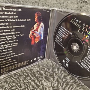 May include: A CD case with a photo of John Denver on the front cover. The case is open, revealing a CD with a picture of John Denver on it. The CD is titled "The Best of John Denver Live".