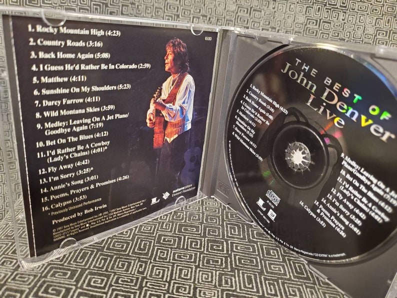 May include: A CD case with a photo of John Denver on the front cover. The case is open, revealing a CD with a picture of John Denver on it. The CD is titled "The Best of John Denver Live".