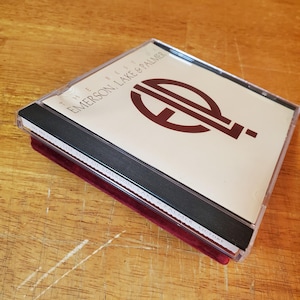 May include: A CD case with the text "THE BEST OF EMERSON, LAKE & PALMER" in black and burgundy. The CD case is clear plastic with a black spine and is resting on a burgundy velvet surface.