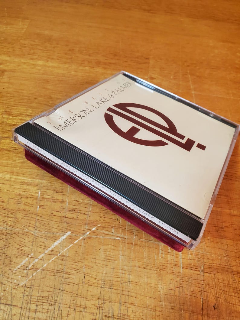 May include: A CD case with the text "THE BEST OF EMERSON, LAKE & PALMER" in black and burgundy. The CD case is clear plastic with a black spine and is resting on a burgundy velvet surface.