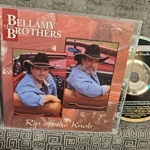 May include: A CD case with a red, white, and blue color scheme. The front cover features a photo of two men in cowboy hats, sitting in a red car. The title of the album is "The Bellamy Brothers: Rip Off The Knob".