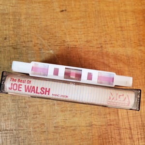K&ouml;nnte beinhalten: Eine Vintage-Kassette mit der Aufschrift "The Best Of JOE WALSH" und ein wei&szlig;-rosa Kassettenrekorder. Das Band ist schwarz mit einem transparenten Fenster, und der Text ist rot. Der Kassettenrekorder ist wei&szlig; mit rosa Akzenten.