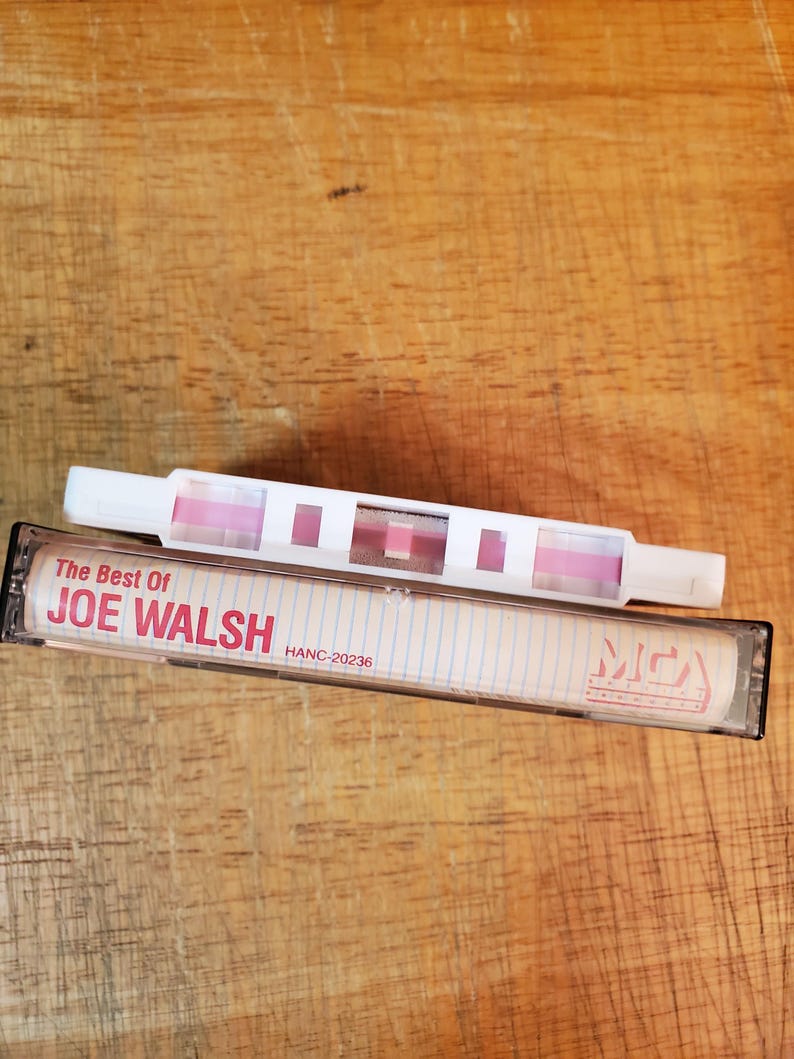 K&ouml;nnte beinhalten: Eine Vintage-Kassette mit der Aufschrift "The Best Of JOE WALSH" und ein wei&szlig;-rosa Kassettenrekorder. Das Band ist schwarz mit einem transparenten Fenster, und der Text ist rot. Der Kassettenrekorder ist wei&szlig; mit rosa Akzenten.