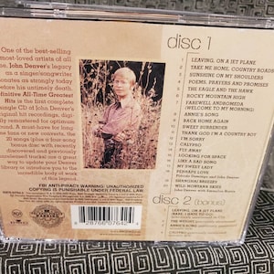 May include: The back cover of a John Denver Definitive All-Time Greatest Hits CD case. It features a photograph of the artist, a tracklist for two discs, descriptive text, a barcode, and an FBI anti-piracy warning logo.