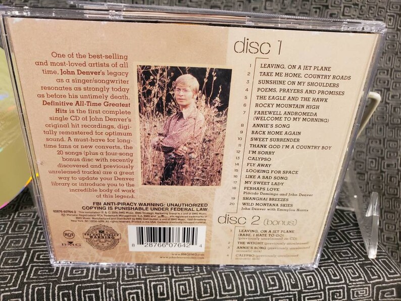 May include: The back cover of a John Denver Definitive All-Time Greatest Hits CD case. It features a photograph of the artist, a tracklist for two discs, descriptive text, a barcode, and an FBI anti-piracy warning logo.