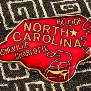 Puede incluir: Un pin de esmalte rojo con forma del estado de Carolina del Norte con las palabras "North Carolina" y "Raleigh" en letras doradas. El pin tambi&eacute;n incluye las ciudades de Asheville y Charlotte en letras doradas.