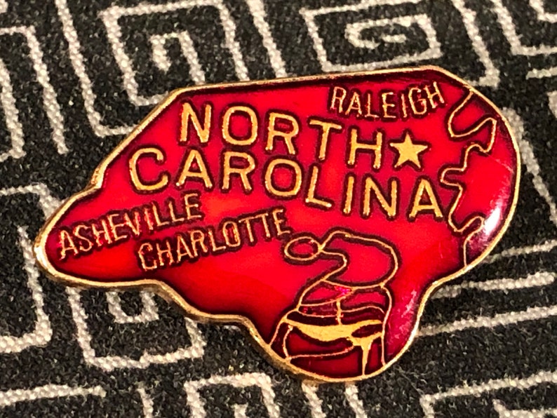 Puede incluir: Un pin de esmalte rojo con forma del estado de Carolina del Norte con las palabras "North Carolina" y "Raleigh" en letras doradas. El pin tambi&eacute;n incluye las ciudades de Asheville y Charlotte en letras doradas.