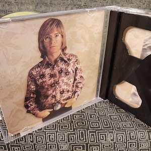 May include: A clear plastic CD jewel case featuring an insert with a photograph of a person with shoulder-length hair and glasses, wearing a long-sleeved shirt with a dark floral pattern and a large belt buckle, set against a beige background.