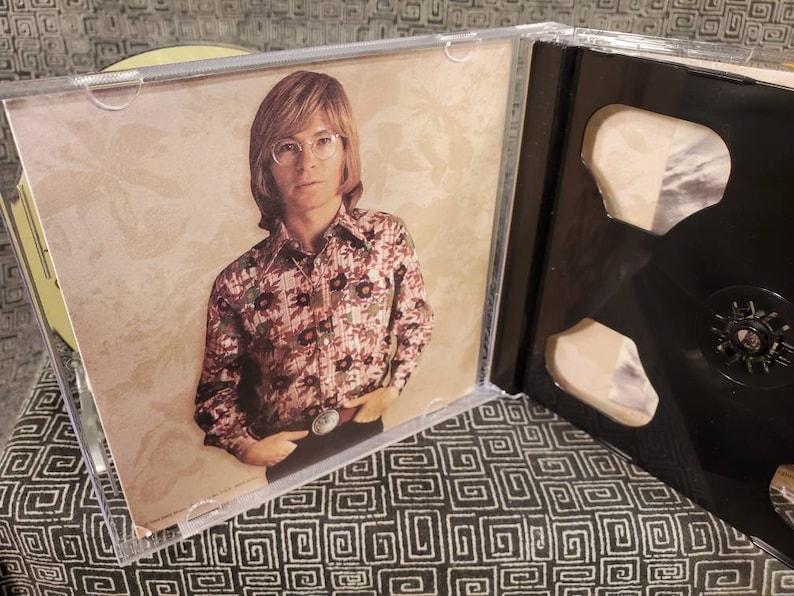 May include: A clear plastic CD jewel case featuring an insert with a photograph of a person with shoulder-length hair and glasses, wearing a long-sleeved shirt with a dark floral pattern and a large belt buckle, set against a beige background.