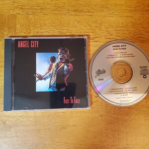 May include: A compact disc and its case are displayed. The CD case features the text "ANGEL CITY FACE TO FACE" with an image of a person reaching out. The CD itself has the text "ANGEL CITY FACE TO FACE" and a list of songs.