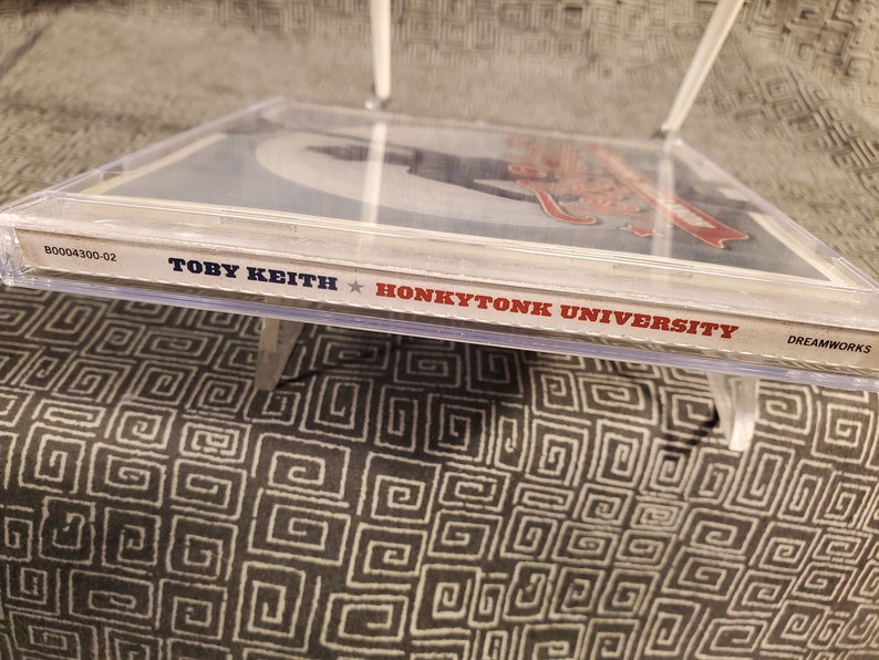 Może przedstawiać: Zbliżenie na pudełko CD z białym tłem i czerwonym tekstem. Tekst brzmi "Toby Keith Honkytonk University" i "Dreamworks".