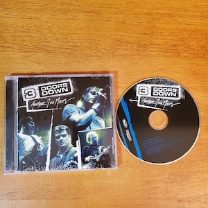 May include: A CD and its case are displayed on a wooden surface. The CD case features the band name "3 Doors Down" and the album title "Another 700 Miles." The CD itself is black with the band's logo and album title.